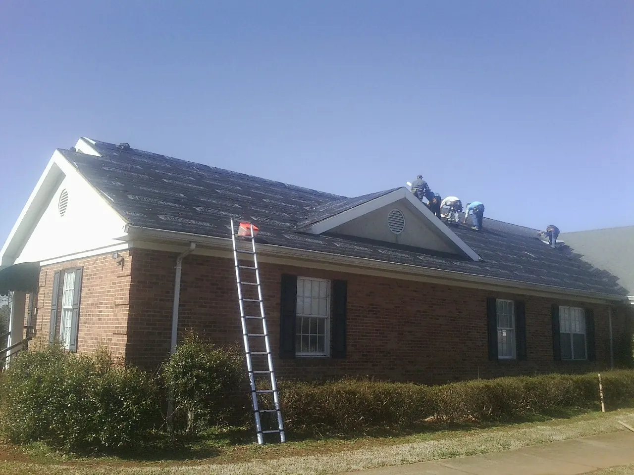 Crew tearing shingles off a brick ranch roof with a ladder set — Metal Roof Repair work by our crew in Calera